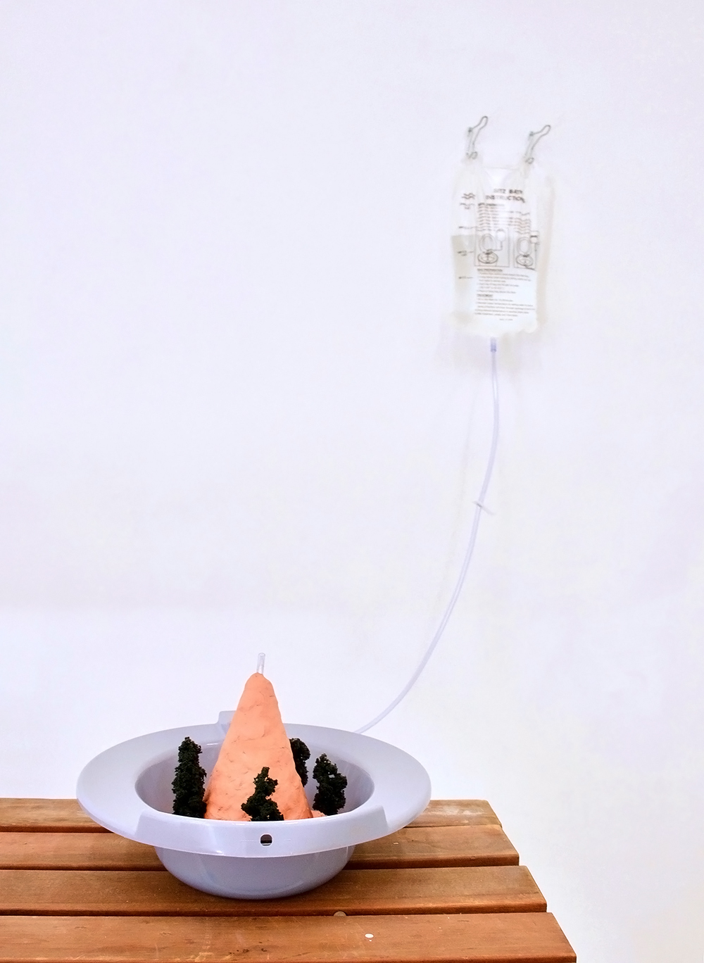 Sculpture of a clay mountain rising out of a sitz bath with a liquid pouch attached to the wall nearby